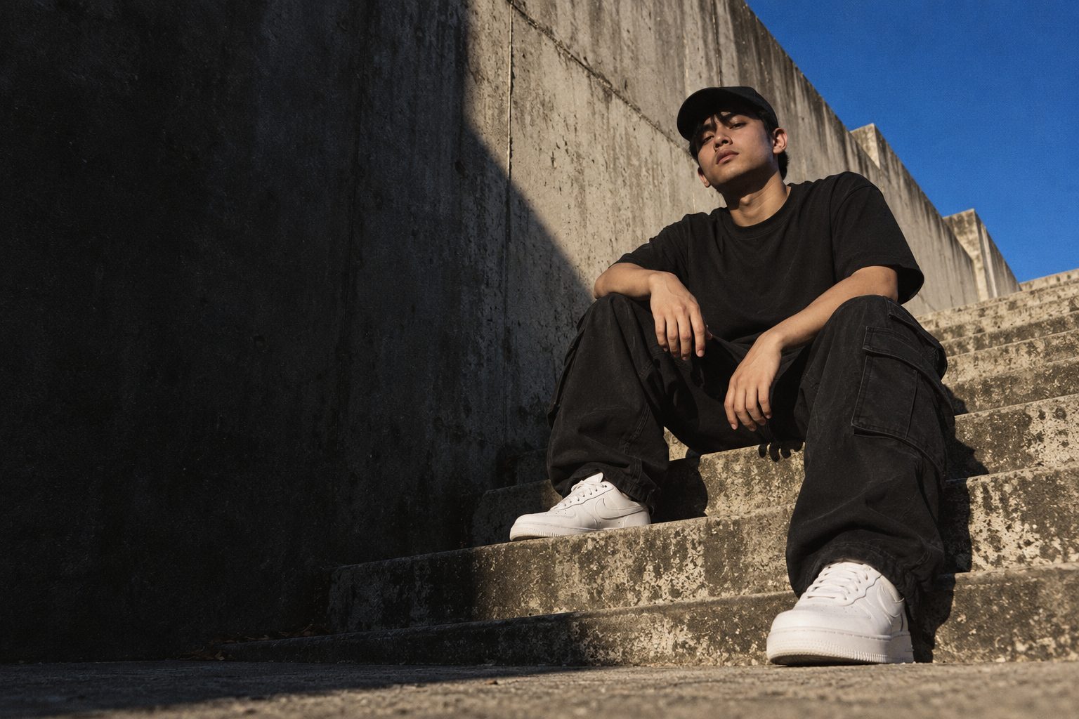 A young man in an oversized black tee and cap sitting on sunlit concrete steps
