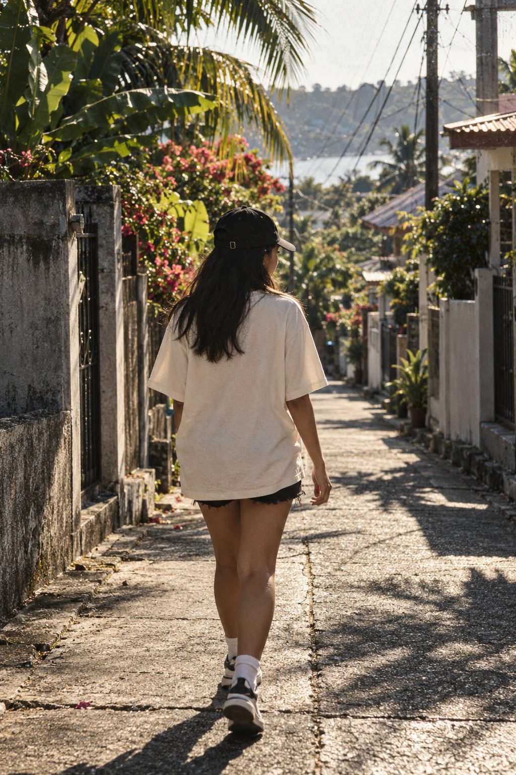 A young woman in an oversized cream tee and black cap walking down a sunny provincial street